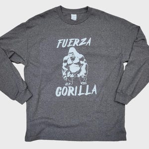 May include: A dark gray long-sleeve shirt featuring a white gorilla graphic. The text "FUERZA GORILLA" is printed above and below the gorilla illustration. The shirt is made of a soft material.