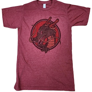 Op de afbeelding: Bordeauxrood t-shirt met een cartoon drakenontwerp in een rode cirkel. De draak is bruin met rode accenten en een lange, krullende snor. Het shirt is gemaakt van een zacht, gemêleerd materiaal.