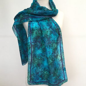 Könnte beinhalten: Ein türkisfarbener Seidenschal mit einem sich wiederholenden Muster aus stilisierten Hunden in einem dunkleren Blau. Der Schal hat einen violetten Saum.