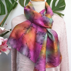 Könnte beinhalten: Ein lebendiger Seidenschal mit Batik-Effekt, der eine Mischung aus Rosa, Orange und Grüntönen aufweist. Der Schal ist über einer Schaufensterpuppe drapiert und zeigt seinen fließenden Stoff und das farbenfrohe Design. Die Abmessungen des Schals sind nicht sichtbar.