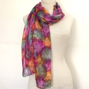 Könnte beinhalten: Ein farbenfroher Schal mit Batikmuster in Rosa-, Lila-, Orange- und Grüntönen. Der Schal ist über einer Schaufensterpuppe drapiert und zeigt seinen fließenden Stoff und sein lebendiges Design. Die Kanten des Schals sind mit dunklem Besatz versehen.