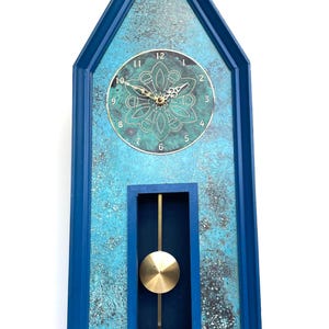 May include: A blue and turquoise wall clock with a pendulum. The clock has a rectangular shape with a pointed top. The clock face is gold with black numbers. The pendulum is gold.