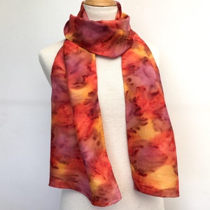 Può includere: Una lunga e sottile sciarpa di seta con un design dipinto a mano in tonalità di arancione, rosso e viola. La sciarpa è legata attorno al collo di un manichino bianco.