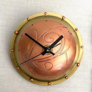 May include: A copper and brass clock with a decorative design and black hands. The clock is round and has a raised rim.