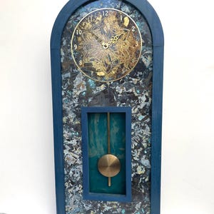 May include: A decorative wall clock with a dark blue frame and a textured background. The clock face is gold with black numerals and ornate detailing. A brass pendulum hangs below. The clock measures approximately 61 cm tall.