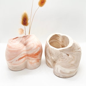 Könnte beinhalten: Zwei Keramikvasen in Form eines menschlichen Körpers mit Marmormuster. Eine Vase ist weiß mit braunen Marmorierungen und die andere ist rosa mit orangefarbenen Marmorierungen. Beide Vasen haben eine runde Öffnung oben. Ein Strauß getrockneter brauner Gräser befindet sich in der rosafarbenen Vase.