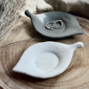May include: Two leaf-shaped ceramic dishes, one white and one gray, with a ring on the gray dish. The dishes are on a wooden surface.