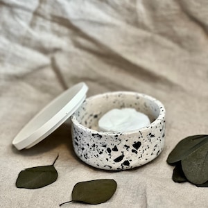 Könnte beinhalten: Ein weißer Keramikbehälter mit einem schwarzen, gesprenkelten Muster und einem passenden Deckel. Der Behälter ist offen und enthält ein weißes, rundes Objekt. Der Behälter steht auf einer beigefarbenen Stoffoberfläche mit verstreuten grünen Blättern.
