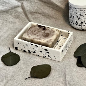 May include: A white soap dish with black speckles, featuring a bar of brown and white swirl soap. The dish has a raised section for draining water.