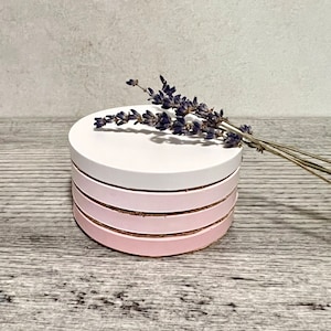 May include: A set of four round coasters with a white top and pink bottom. The coasters are stacked on top of each other. A sprig of lavender is resting on top of the coasters.