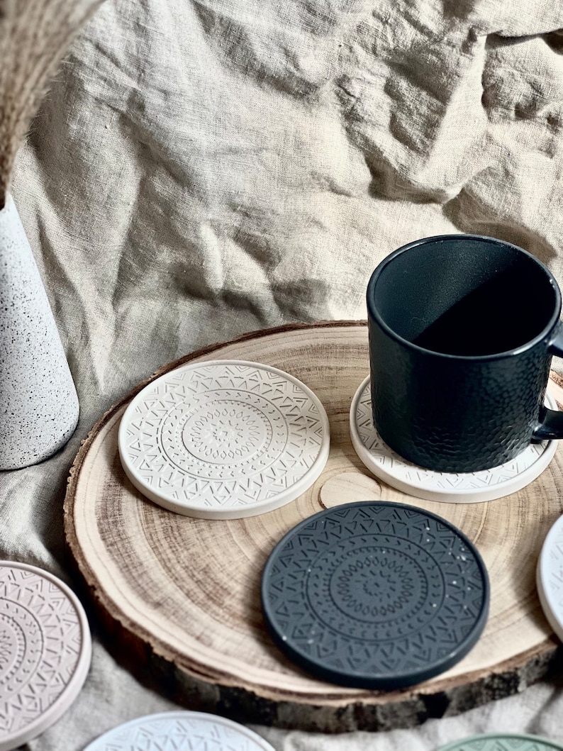 Puede incluir: Conjunto de cuatro posavasos redondos con un patr&oacute;n geom&eacute;trico. Los posavasos est&aacute;n hechos de un material gris claro y gris oscuro. Los posavasos est&aacute;n sobre una superficie de madera con una taza negra.