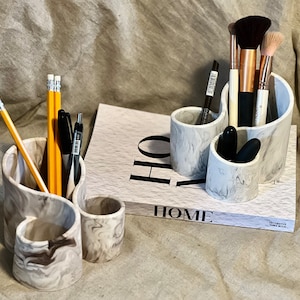 May include: Two white marble swirl ceramic desk organizers. One holds pencils and pens, the other holds makeup brushes. Both are on a beige fabric surface with a book that says "HOME" on the cover.