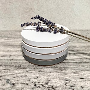 May include: Set of four white coasters with a grey and gold stripe design. The coasters are stacked on top of each other and a sprig of lavender is resting on top.