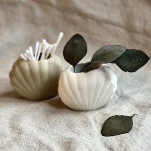 May include: Two ceramic shell-shaped vases, one beige and one white, with dried eucalyptus leaves. The beige vase is filled with cotton swabs.