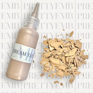 May include: A clear glass bottle with a brown liquid inside. The bottle has a white label that says "CREAM TAN". The bottle is sitting on a white surface with a pile of broken tan-colored pieces of something.