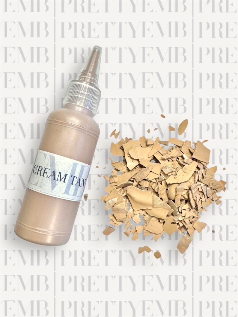 May include: A clear glass bottle with a brown liquid inside. The bottle has a white label that says "CREAM TAN". The bottle is sitting on a white surface with a pile of broken tan-colored pieces of something.