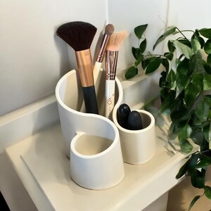 May include: White ceramic makeup brush holder with three compartments. The holder has a wavy, organic design. It is holding three makeup brushes and two black makeup applicators.