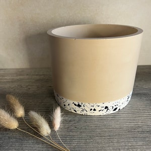 May include: A beige ceramic planter with a white and black terrazzo design around the top edge. The planter is sitting on a wooden surface.