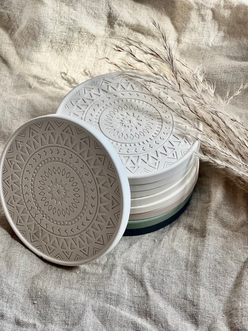 Puede incluir: Conjunto de seis posavasos redondos de color beige con un patr&oacute;n geom&eacute;trico. Los posavasos est&aacute;n apilados uno encima del otro.
