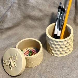 May include: Two small beige concrete containers with a honeycomb pattern. One container has a lid with a bee design and the other container has pencils and pens inside.