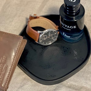 May include: A black tray holds a brown leather wallet, a wristwatch with a brown leather band, and a bottle of Dior Sauvage Eau de Parfum. The watch has a silver case and a black face.