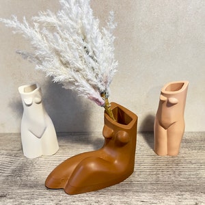 May include: Three ceramic vases shaped like the human form. The vases are in white, light pink, and brown. A bouquet of dried white pampas grass is in the brown vase.