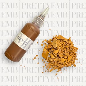 May include: A brown glass bottle with a white label that says "TOFFEE" and a clear plastic dropper top. The bottle is sitting on a white surface with a pile of broken, orange-brown pieces of toffee.