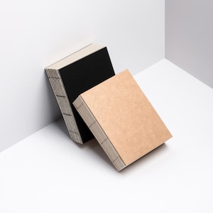 以下が含まれることがあります： 白角に寄りかかる、黒とタンの2冊の長方形ノート。ノートは背表紙にステッチが見え、ページが積み重なっています。タン色のノートが黒色のノートの前にあります。