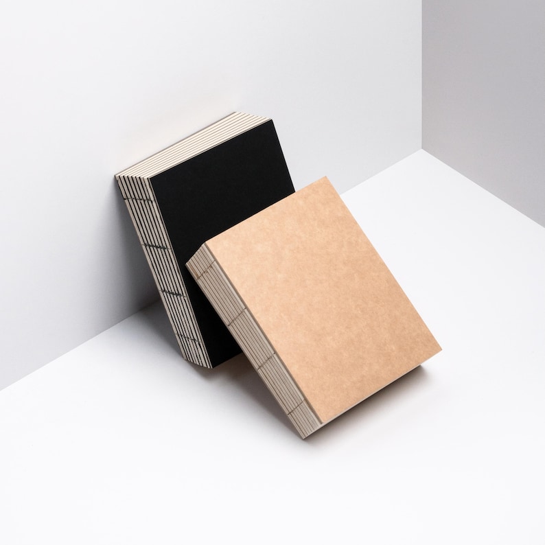 Peut inclure: Deux cahiers rectangulaires, un noir et un beige, sont appuy&eacute;s contre un coin blanc. Les cahiers ont des coutures visibles le long du dos et une pile de pages. Le cahier beige est devant le noir.