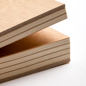 May include: Two stacks of rectangular, light brown paper pads are shown against a white background. The pads are made of thick paper, with visible edges. The top pad is slightly open, revealing the pages within. The paper has a natural, textured appearance.