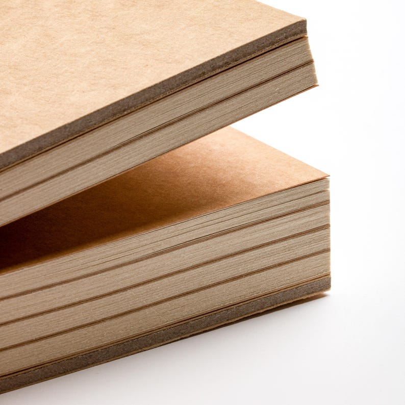 May include: Two stacks of rectangular, light brown paper pads are shown against a white background. The pads are made of thick paper, with visible edges. The top pad is slightly open, revealing the pages within. The paper has a natural, textured appearance.