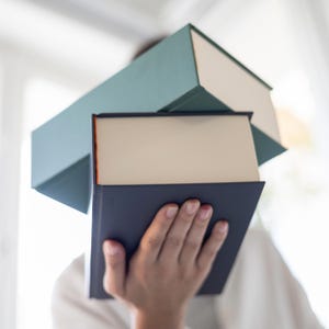 May include: A person is holding three books. The top book is teal green, the middle book is white, and the bottom book is dark blue.
