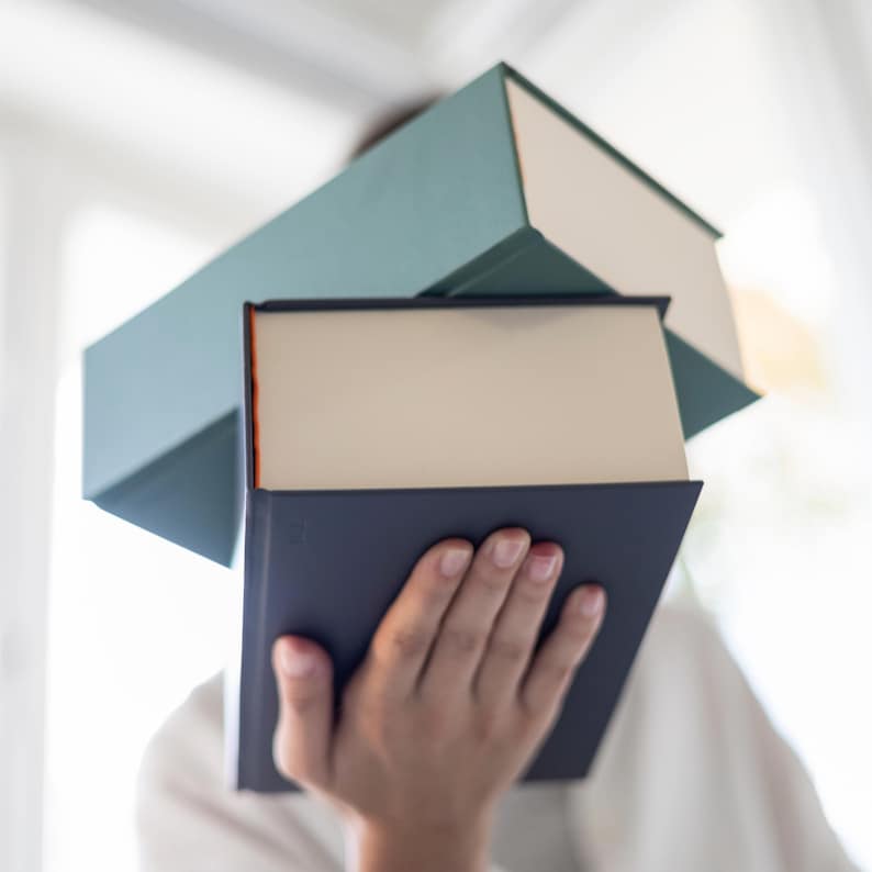 May include: A person is holding three books. The top book is teal green, the middle book is white, and the bottom book is dark blue.