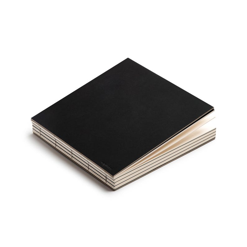 May include: A black notebook with a textured cover, partially open to reveal the cream-colored pages. The binding is visible, with a series of stitched lines. The notebook is set against a white background.