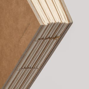 May include: Close-up of a handmade journal with a brown cover and cream-colored pages. The pages are sewn together with visible stitching along the spine. The cover has the embossed text "less notes".