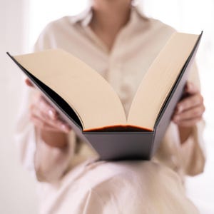 May include: A person is reading a large, open book with a black cover and cream-colored pages. The book is held open with both hands.