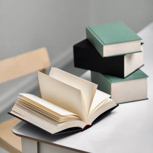 May include: An open book with white pages and a black cover lies on a white table. Three books are stacked on top of each other in the background. The top book is green, the middle book is black, and the bottom book is green.