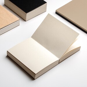 Peut inclure: Un carnet ouvert avec des pages cr&egrave;me, entour&eacute; de plusieurs carnets ferm&eacute;s. Les carnets ferm&eacute;s ont des couvertures noires, beiges et marron clair. Les pages du carnet ouvert sont l&eacute;g&egrave;rement blanc cass&eacute;, et les bords des pages sont visibles.