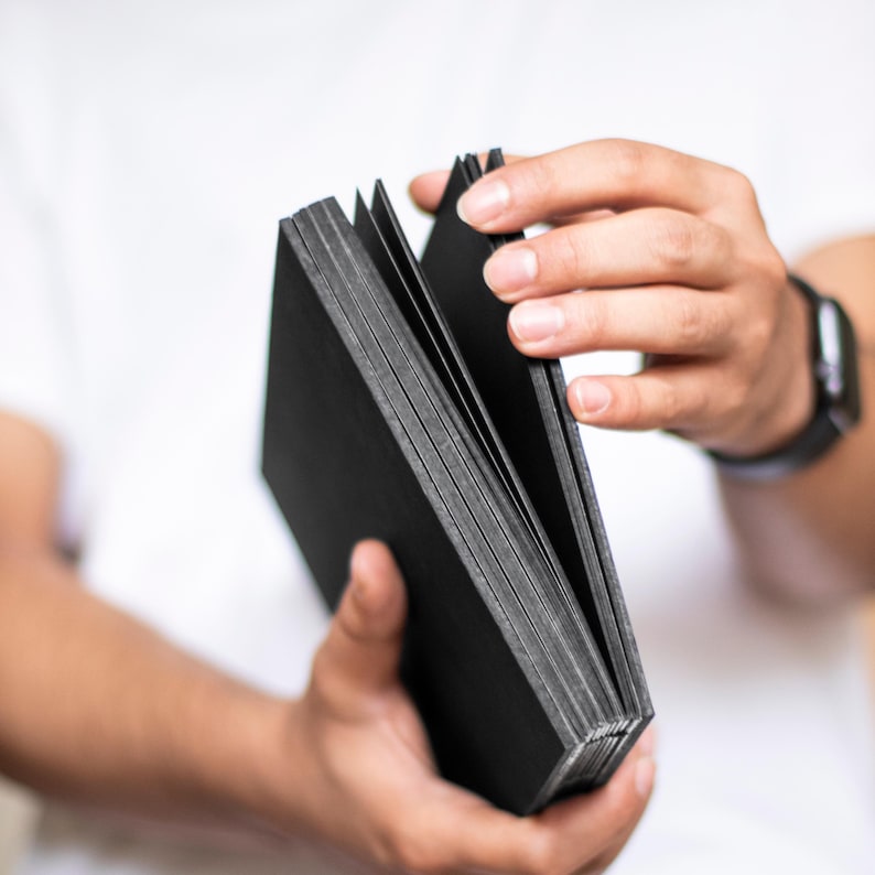 May include: A black, leather-bound journal with a textured cover. The journal is open and fanned out, showing the pages inside.