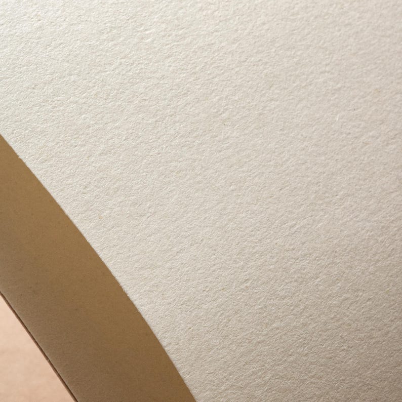 May include: Close-up of a textured, off-white paper surface with a diagonal strip of light brown material. The paper has a slightly rough, matte finish, and the lighting creates subtle shadows.