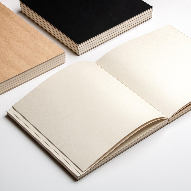 May include: An open notebook with cream-colored pages, alongside two closed notebooks. One notebook has a black cover, and the other has a light brown cover. The pages have a smooth texture, and the edges of the pages are visible.