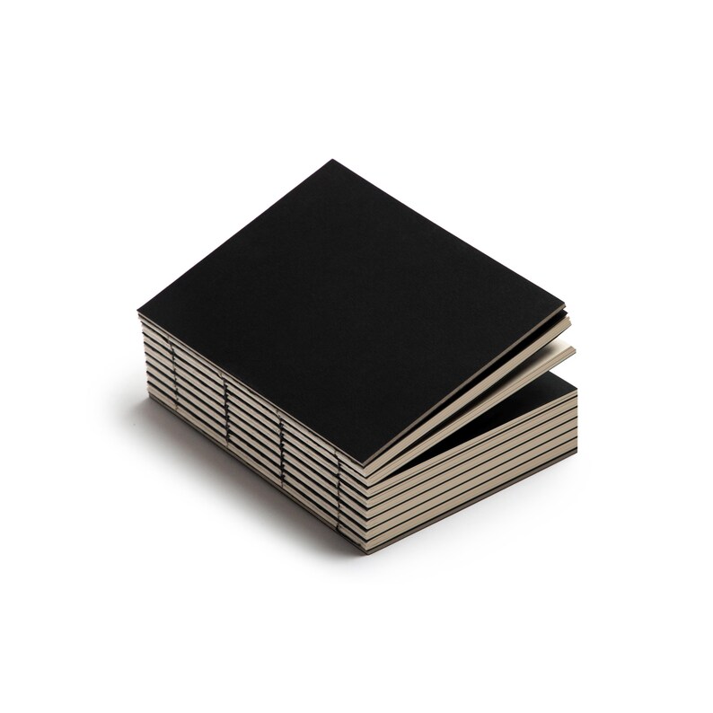 Peut inclure: Une pile de petits cahiers noirs avec des pages cr&egrave;me. Les cahiers sont rectangulaires et semblent &ecirc;tre reli&eacute;s ensemble. Le cahier sup&eacute;rieur est l&eacute;g&egrave;rement ouvert, r&eacute;v&eacute;lant les pages int&eacute;rieures. Le fond est blanc uni.