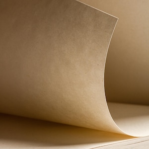 May include: Close-up of a sketchbook with pages made of light brown paper. The top page is slightly curved, revealing the texture and color of the paper. The pages are stacked, suggesting a new or unused notebook.
