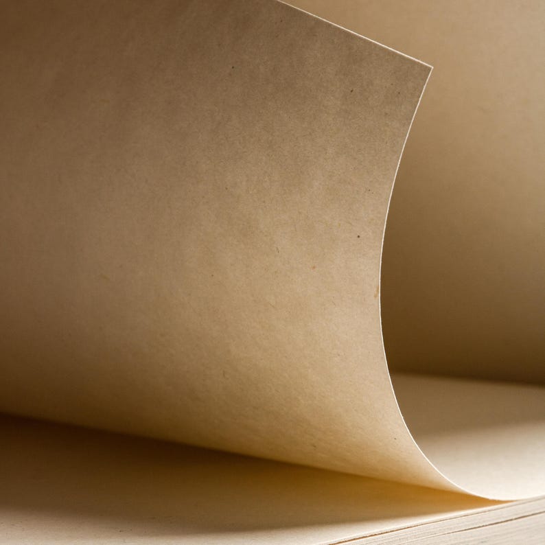 May include: Close-up of a sketchbook with pages made of light brown paper. The top page is slightly curved, revealing the texture and color of the paper. The pages are stacked, suggesting a new or unused notebook.