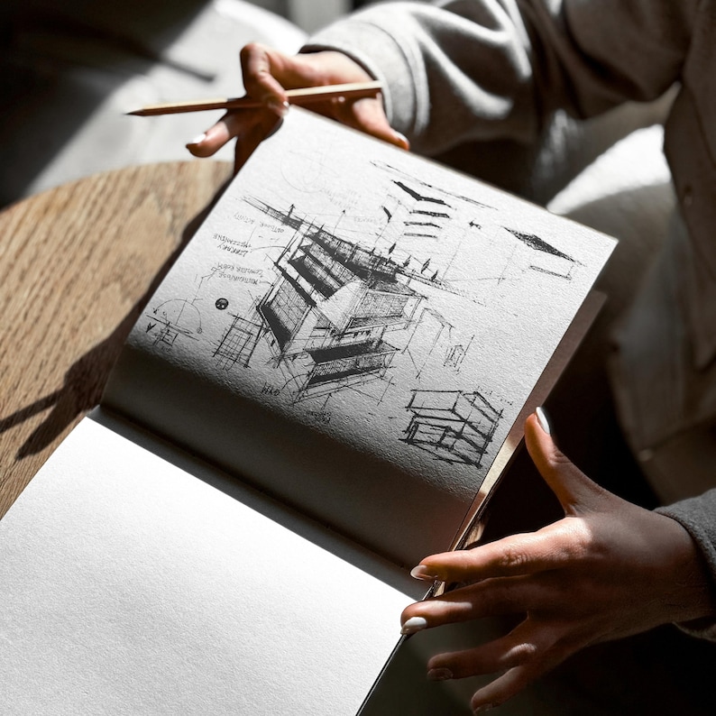 May include: An open sketchbook with architectural sketches in pencil. The drawings depict building designs with detailed lines and shading. A wooden pencil rests on the page, held by a person. The sketchbook is on a wooden table.
