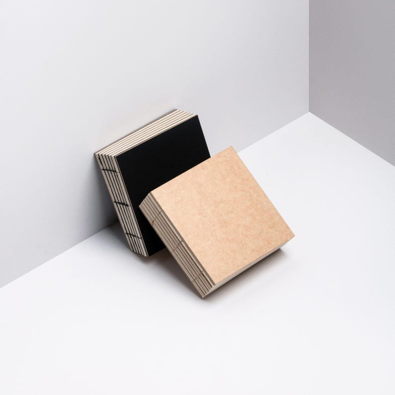May include: Two square notebooks, one black and one tan, are stacked and leaning against a white wall. The pages are visible on the sides, and the binding is a light color. The notebooks are on a white surface.