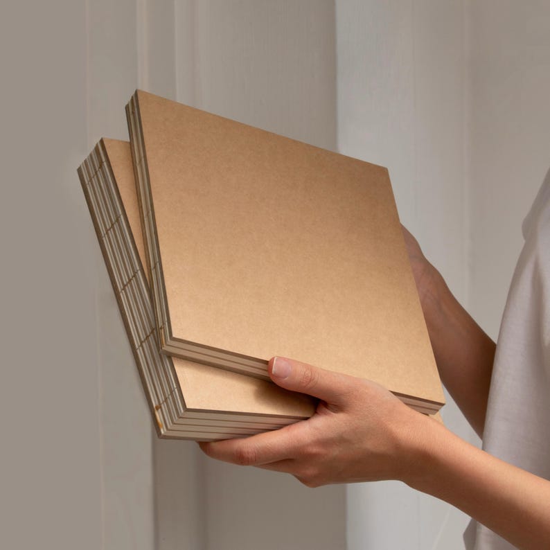 May include: A stack of square, kraft-colored paper notebooks. The notebooks are held in a person's hands, with the edges of the pages visible. The paper has a natural, textured appearance, suggesting a handmade or eco-friendly product.