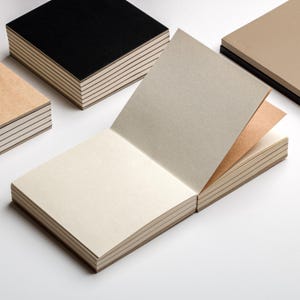 May include: Several square notebooks with different colored covers are displayed. One notebook is open, revealing the interior pages. The covers are black, tan, and beige. The pages are a light beige color.