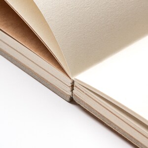 May include: Close-up of an open notebook with cream-colored pages. The pages are slightly textured and bound together with a gray spine. The paper is thick and appears to be high-quality, suitable for writing or sketching.