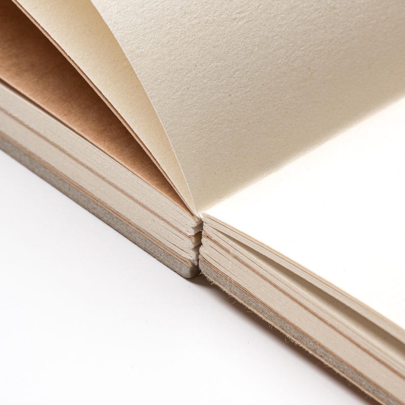 May include: Close-up of an open notebook with cream-colored pages. The pages are slightly textured and bound together with a gray spine. The paper is thick and appears to be high-quality, suitable for writing or sketching.